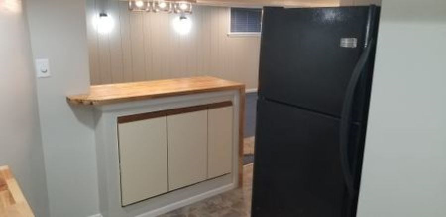 A black refrigerator stands next to a wooden counter with beige cabinets under a strip of lights.