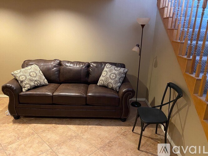 A brown leather couch with two cushions and a black chair beside a lamp.