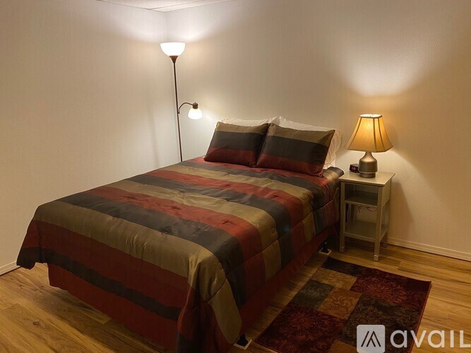 A bed with a striped comforter and two pillows sits next to a small table with a lamp on it.