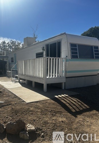A white mobile home with a porch is in a sunny, rocky lot.
