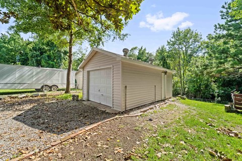 A small house with a garage is surrounded by trees and has a gravel area in front.