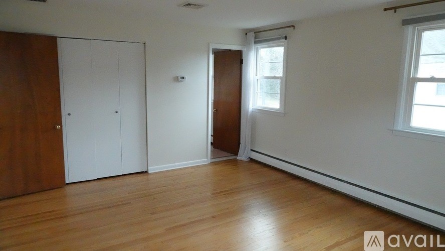 A room with wooden floors, a white door, a window with white curtains, and a wooden wardrobe.