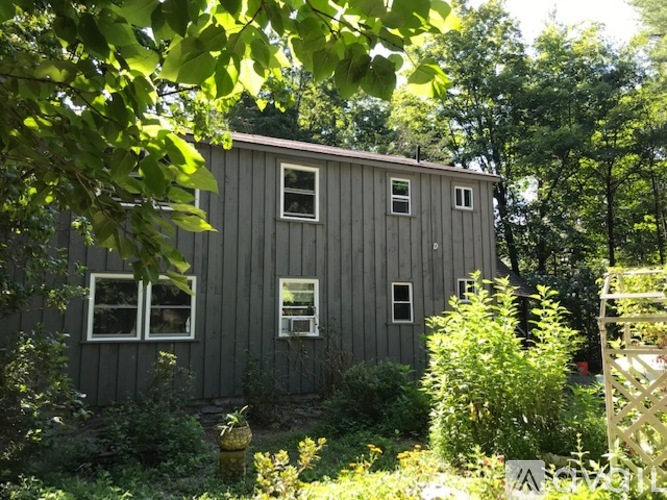 A small grey building with a white window is surrounded by greenery.