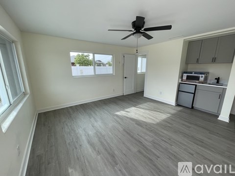 A spacious room with a ceiling fan and light, hardwood flooring, and a window with a view of trees and a house.