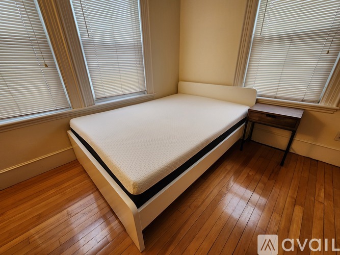 A bed with a white mattress and wooden frame in a room with wooden flooring and windows.