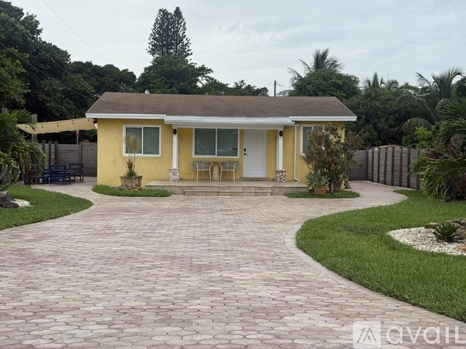 A yellow house with a brown roof and a white door is surrounded by a brick pathway and greenery.