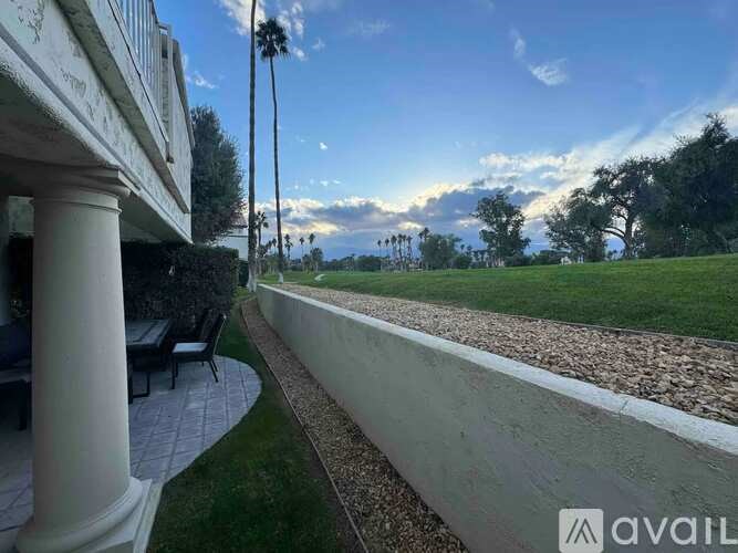 A patio with a white column and a bench is surrounded by a grassy area and trees.