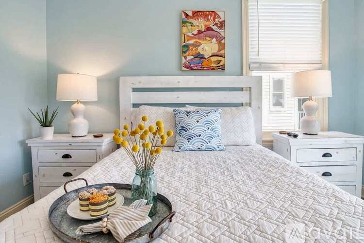 A bedroom with a white bed, a tray with pastries and yellow flowers on it, and a painting above the bed.
