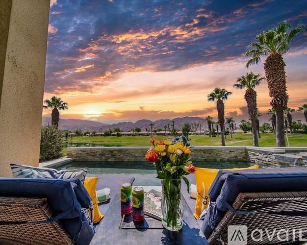 A table with a vase of flowers, a drink, and a plate of food is set up on a patio with a view of palm trees and a sunset.