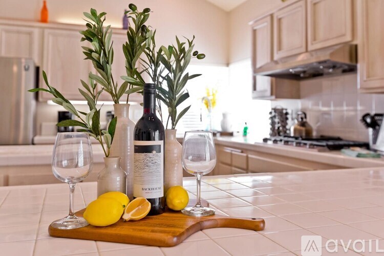 A kitchen counter with a bottle of wine, two glasses, a sprig of rosemary, and a lemon.