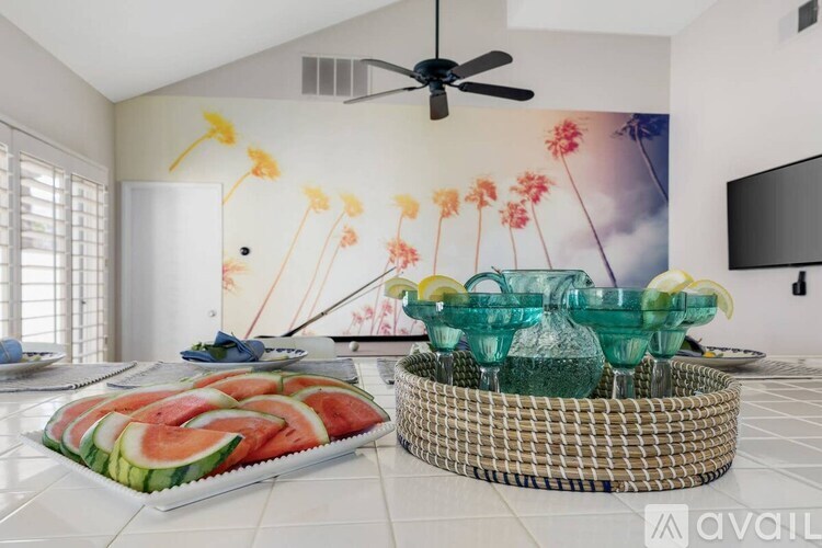 A room with a fan, a wall mural, and a table with a watermelon platter and glasses on it.