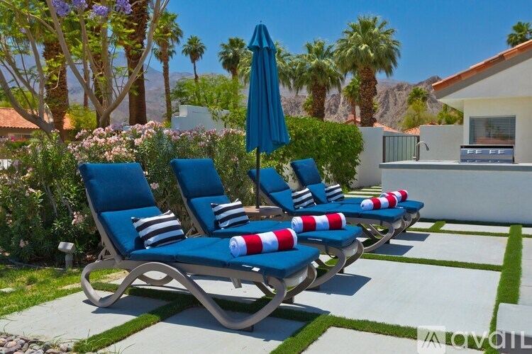 A row of blue lounge chairs with red and white pillows are arranged on a patio.