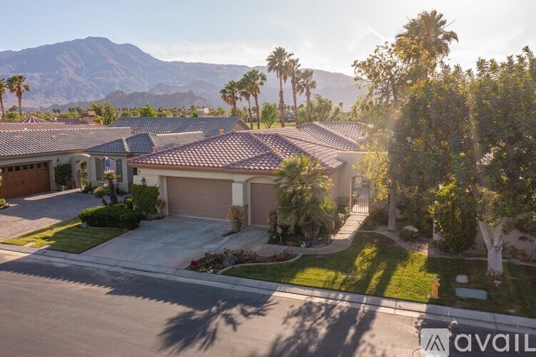 A house with a brown roof and a garage door is surrounded by greenery and palm trees.