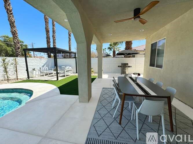 A patio with a table and chairs is surrounded by a pool and palm trees.
