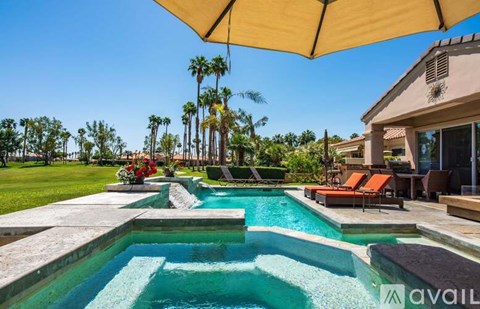 A pool with a waterfall and a patio with chairs and an umbrella.