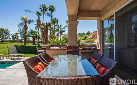A patio with a glass table and chairs with a pool and palm trees in the background.