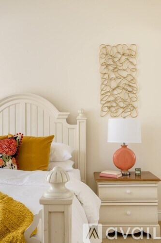 A bedroom with a white bed, a white nightstand with a lamp and a picture on the wall.