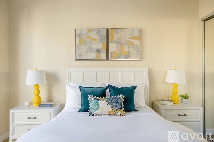 A bedroom with a white bed, two yellow lamps, and a painting above the bed.