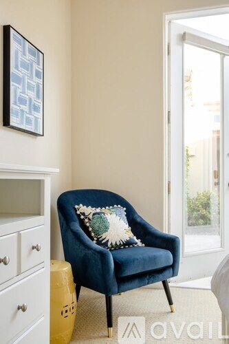A blue chair with a pillow and a white dresser with a painting on the wall.