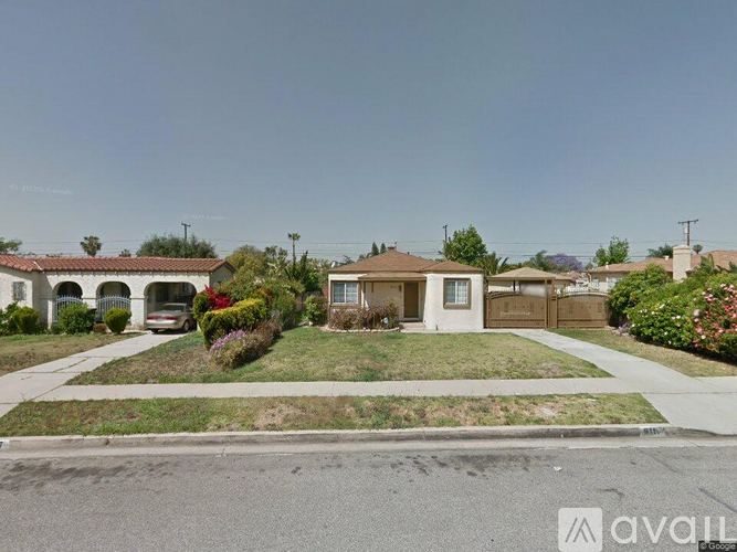 A house with a brown roof and a white wall is surrounded by a green lawn and a sidewalk.