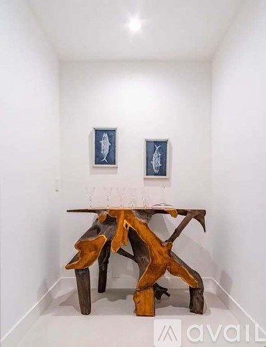 A wooden table with a unique design is in the center of a white room with two framed pictures on the wall.