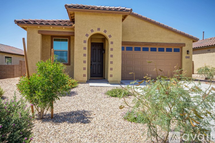 A house with a brown stucco exterior and a black garage door is surrounded by a gravel driveway and small shrubs.