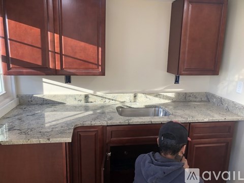 A person in a blue hoodie is standing in a kitchen with brown cabinets and a marble countertop.