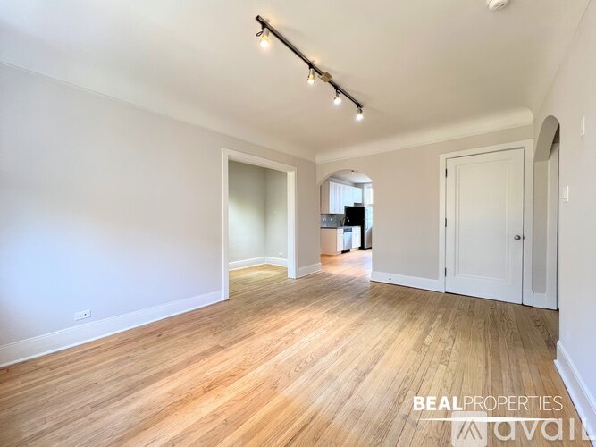 A spacious room with wooden flooring and white walls, with a door on the right and a window on the left.
