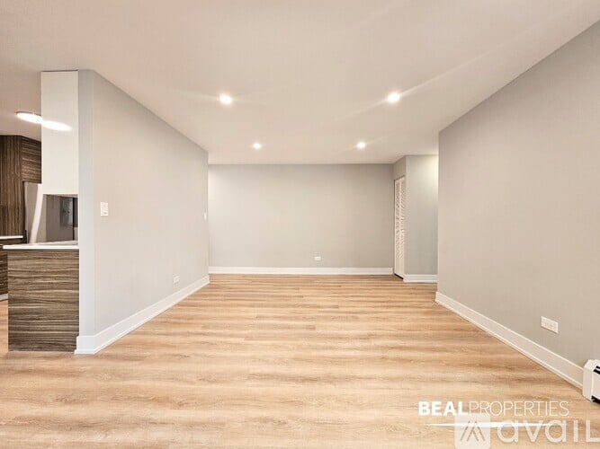 A spacious room with wooden flooring and white walls, with a kitchen area visible on the left.