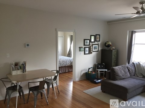 A living room with a grey couch, a white dining table with four chairs, and a bed with a white comforter in the background.