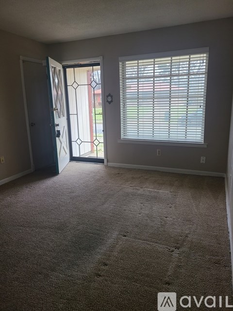 A room with a carpeted floor, a window with blinds, and a door leading to a hallway.
