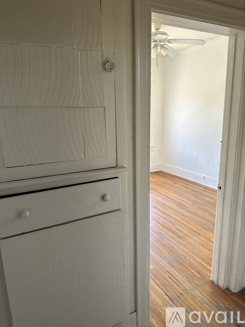 A white cabinet with a drawer is in the foreground of an empty room with wooden flooring and a ceiling fan.