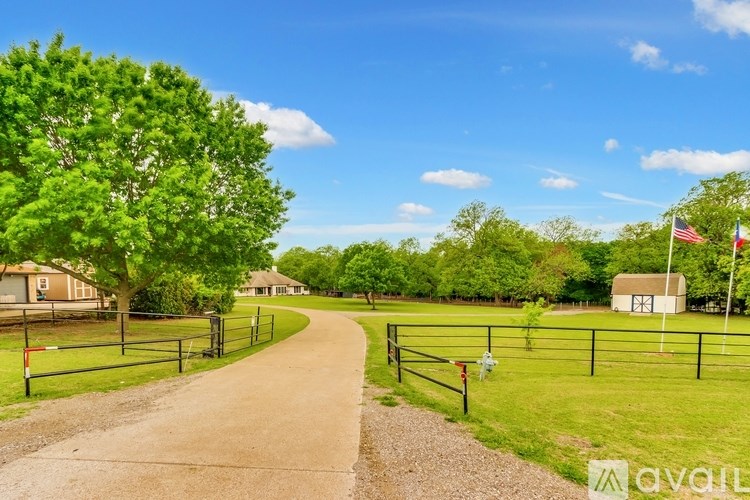 A dirt road leads through a grassy area with a fence on one side and a house on the other.