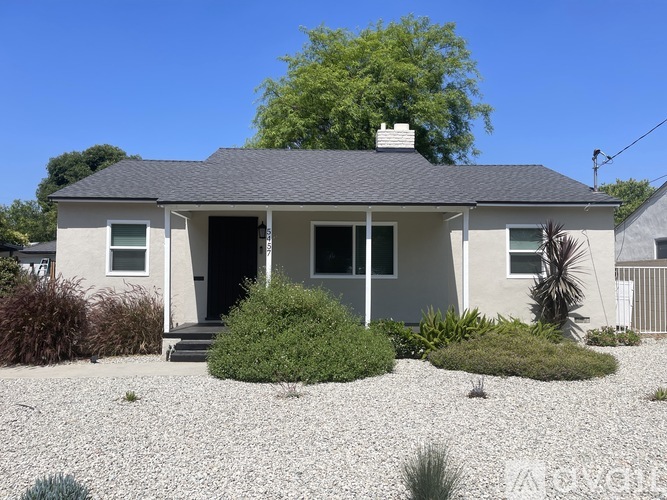 A house with a grey roof and white walls is surrounded by a gravel driveway and small bushes.