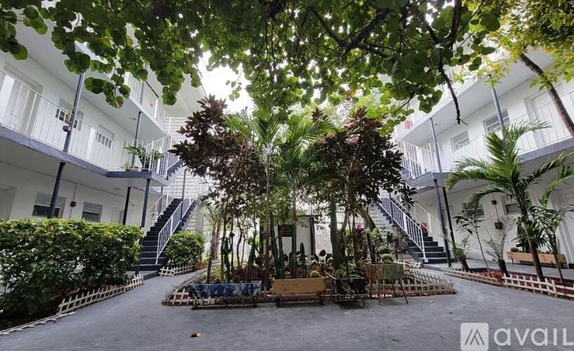 A courtyard with trees and plants in the foreground and white buildings in the background.