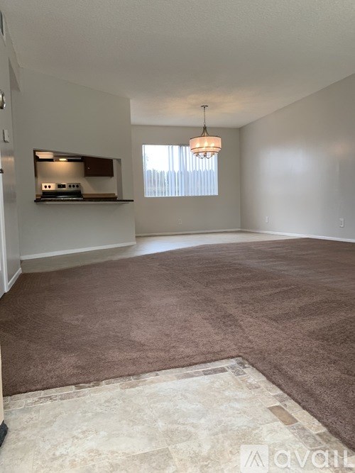 A room with a brown carpet and a kitchen area in the background.