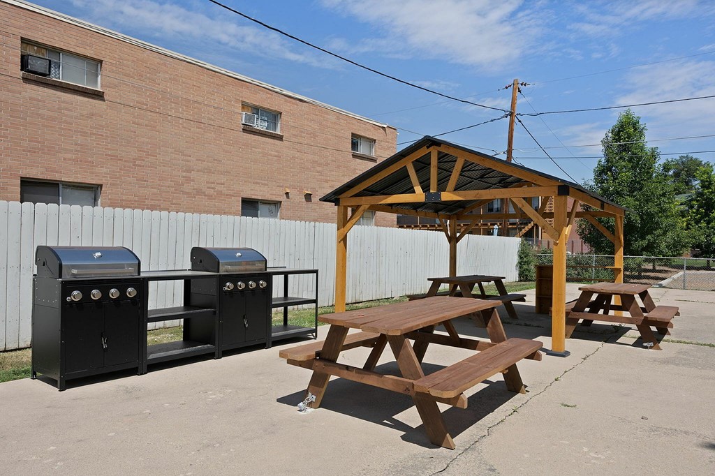 A wooden picnic table with a grill and trash cans in front of a brick building.