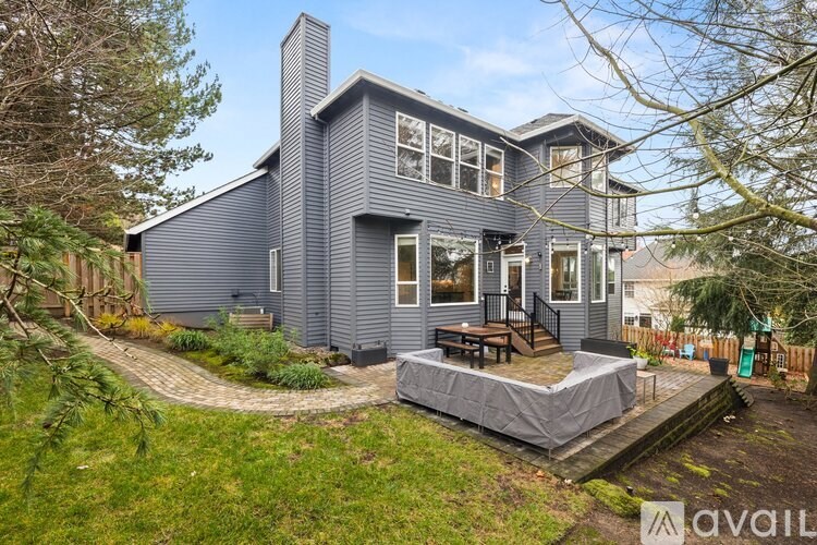 A house with a grey exterior is surrounded by greenery and has a patio with a table and chairs.