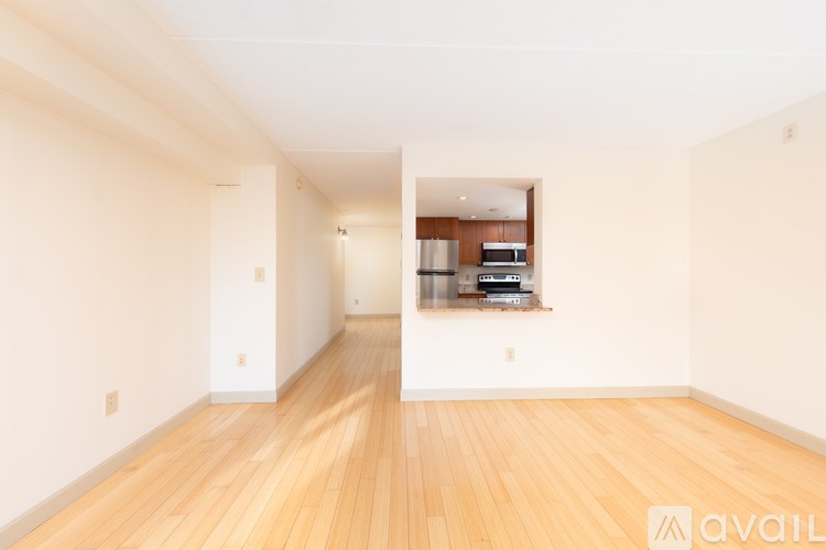 A room with wooden floors and white walls, with a kitchen visible through a window.