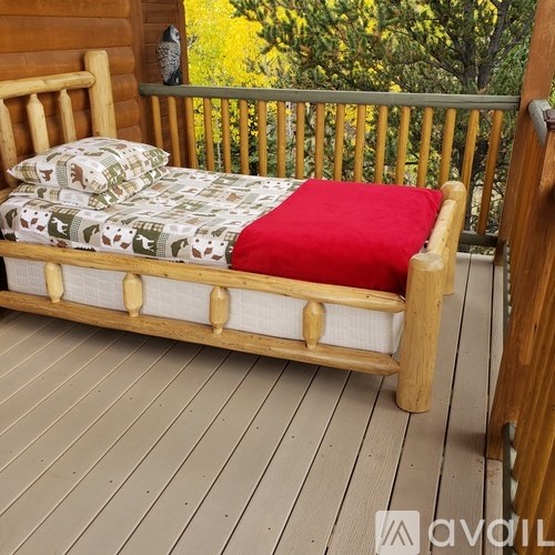 A wooden bed with a red blanket and a green pillow is on a wooden deck.