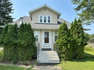 A house with a grey roof and white walls has a small front yard with a few trees.