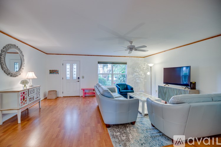 A living room with a white couch, a white coffee table, a white side table, a white wall, a white ceiling, a white door, a white fan, a white rug, a white table lamp, a white wall clock,