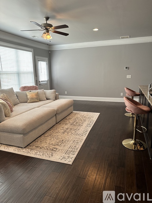 A living room with a beige couch, a rug, a ceiling fan, and a bar area with pink chairs.