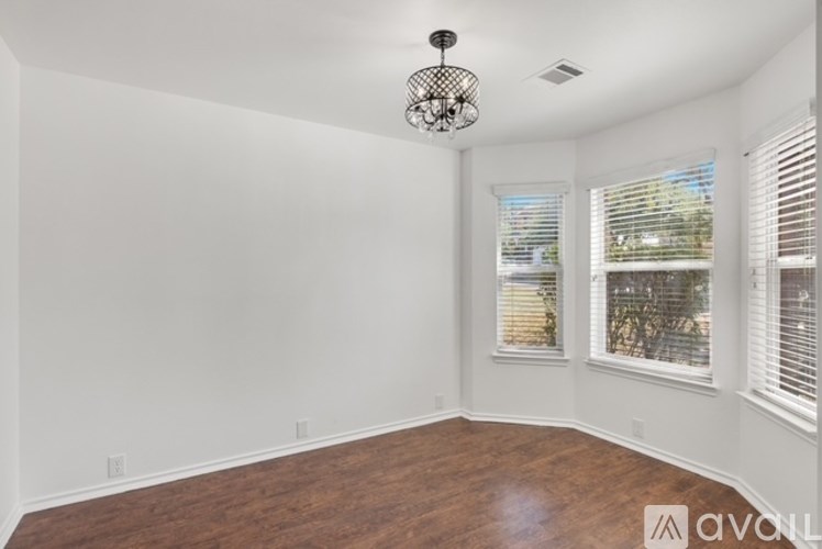 A room with wooden floors and white walls, featuring a chandelier and two windows with blinds.
