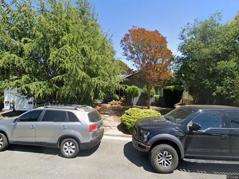 Two cars are parked on a street with trees and a house in the background.