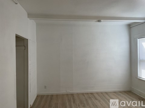 A room with a wooden floor and white walls, with a door on the left and a window on the right.