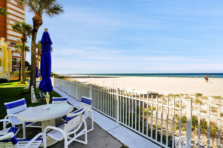 A white table with blue chairs and a blue umbrella is on a patio overlooking the beach.