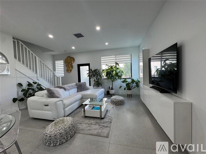 A modern living room with a white sofa set, a glass coffee table, and a flat-screen TV mounted on the wall.