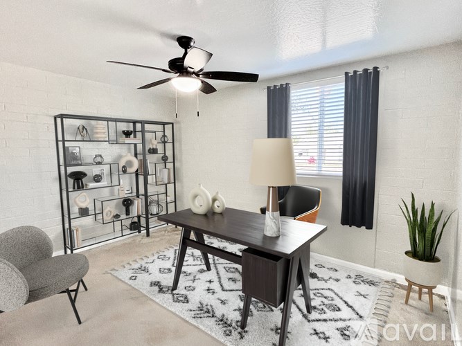 A living room with a black table, a grey chair, a white lamp, a black and white rug, a plant, and a fan.