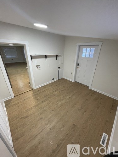 A room with wooden flooring and white walls, with a door on the right and a window on the left.